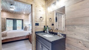 Une chambre dans un chalet en bois, avec un lit, une salle de bain comprenant un lavabo et un miroir, et une fenêtre avec des stores.