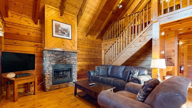 A wooden interior with a stone fireplace, a sofa, a coffee table, a TV, and a staircase. - Adorable log cabin with front-back deck and hot tub (Pigeon Forge)