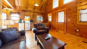 A cozy cabin interior with wooden walls, a sofa, a coffee table, a dining area, and a kitchen. - Adorable log cabin with front-back deck and hot tub (Pigeon Forge)