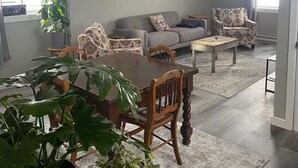 A spacious living room with a dining area, grey sofas, patterned armchairs, and two framed pictures on the wall. - Belmont Stayover (Belmont)