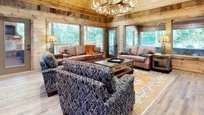 A spacious living room with wooden walls and ceiling, a large chandelier, and a view of the outdoors through large windows. - Elegant Lodge, for 12・Pool・Hot Tub・Theater・Games (Sevierville)