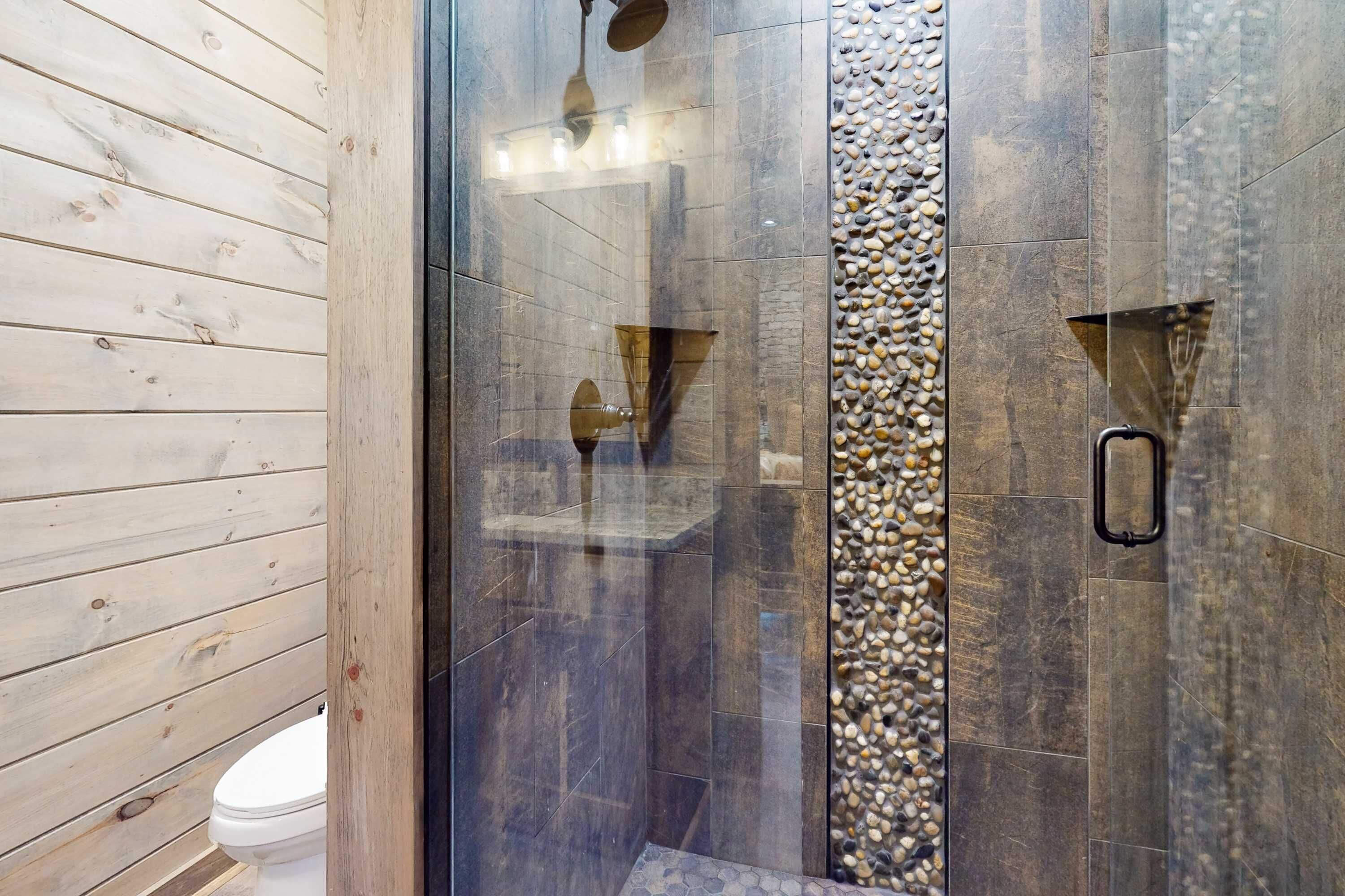 A modern bathroom with a wooden wall, a glass shower enclosure, and a pebble accent wall.