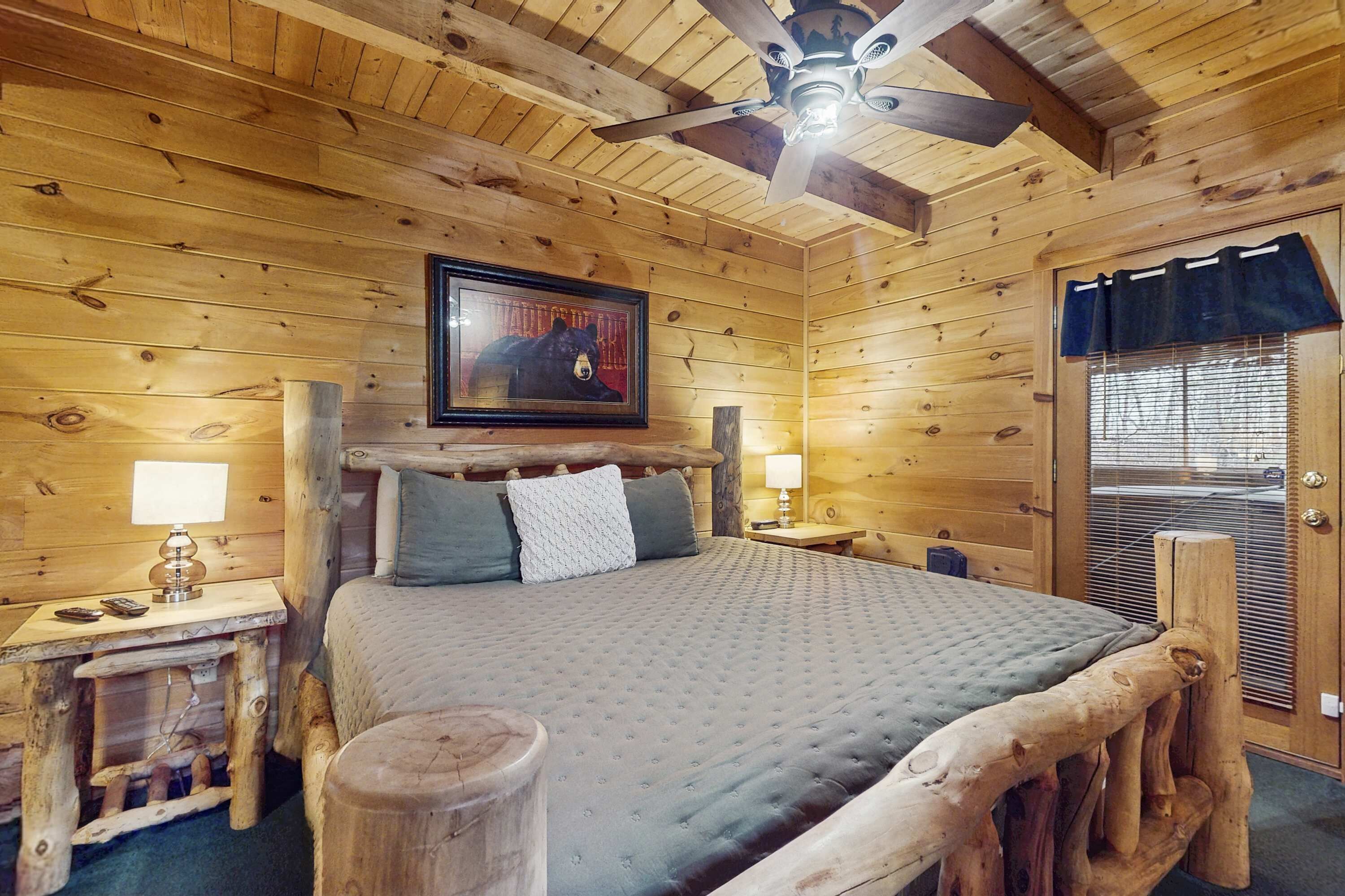 Un dormitorio de cabaña de madera con cama, cabecera de madera, ventilador de techo, un cuadro en la pared y una ventana con persianas.