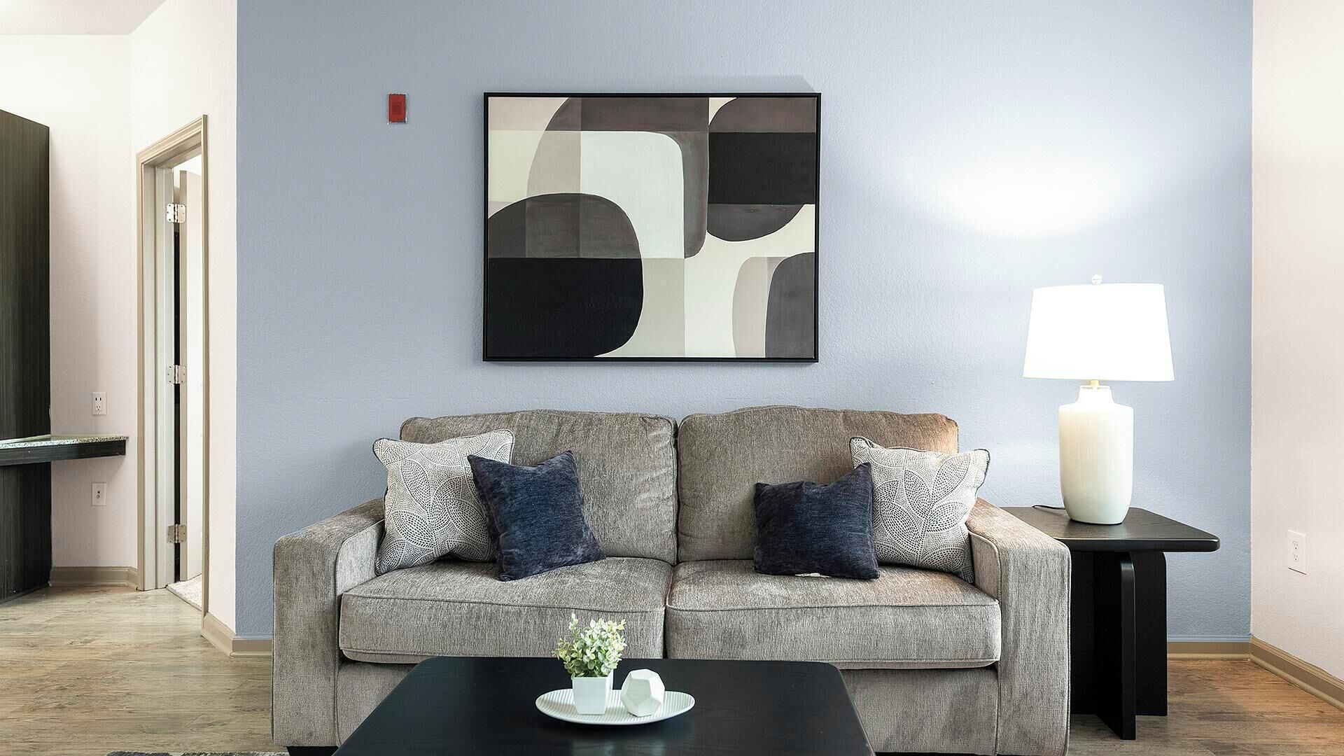 Una sala de estar con un sofá gris, dos almohadas, una mesa de centro negra con un jarrón blanco y un cuadro abstracto enmarcado en la pared.