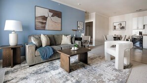 A modern living room with a grey sofa, a coffee table, and a white armchair. There is a painting on the wall and a kitchen in the background. - Landing | Chic 2BD, Gym, Clubhouse (Concord)