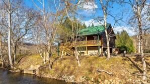 Une cabane dotée d’une terrasse en bois et d’un toit végétalisé, entourée d’arbres et bordée par une rivière.