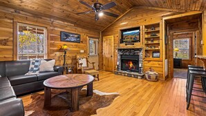 L’intérieur chaleureux d’un chalet en bois comprend une cheminée, un canapé en cuir, une table à manger en bois et un ventilateur de plafond.