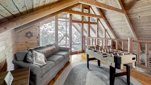 Un appartement-loft chaleureux avec un plafond en bois, un canapé gris, une table de baby-foot et de grandes fenêtres donnant sur la verdure.