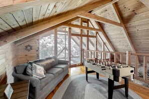 Un loft acollidor amb sostre de fusta, un sofà gris, una taula de futbolí i grans finestres que donen vistes a arbres.