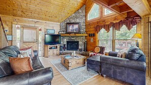 Interior acogedor de una cabaña de troncos con chimenea de piedra, sofás de cuero, televisor y un amplio ventanal.