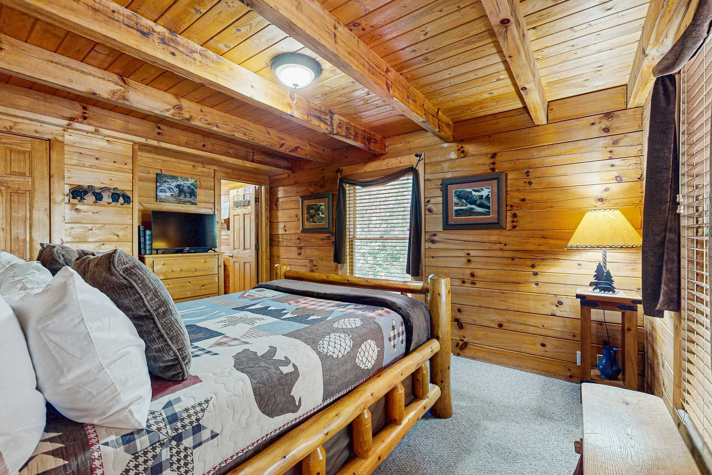 A wooden cabin room with a bed, a TV, a window with blinds, and a lamp.