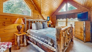 Un dormitorio rústico de madera con una cama grande, televisión y vistas al bosque a través de las ventanas.