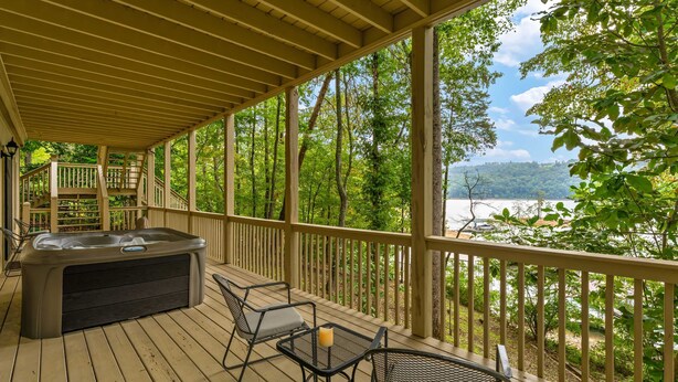 Eine überdachte Terrasse mit Whirlpool, Gartenmöbeln und Blick auf einen von Bäumen umgebenen See.