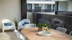 Un comedor moderno con una mesa de madera y sillas blancas, una planta en maceta y una barandilla de cristal con vistas a un edificio.