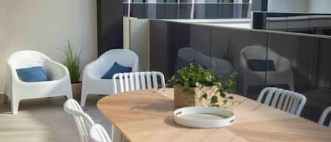 A modern dining area with a wooden table and white chairs, a potted plant, and a glass railing overlooking a building.