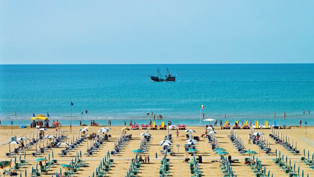Una playa con filas de tumbonas y sombrillas, un mar azul cristalino y un barco pesquero a lo lejos.