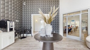 Ett modernt vardagsrum med en vägg dekorerad med ett geometriskt mönster, en kaffemaskin, ett runt bord med en vas med pampasgräs och en skjutdörr i glas som leder till en matplats.
