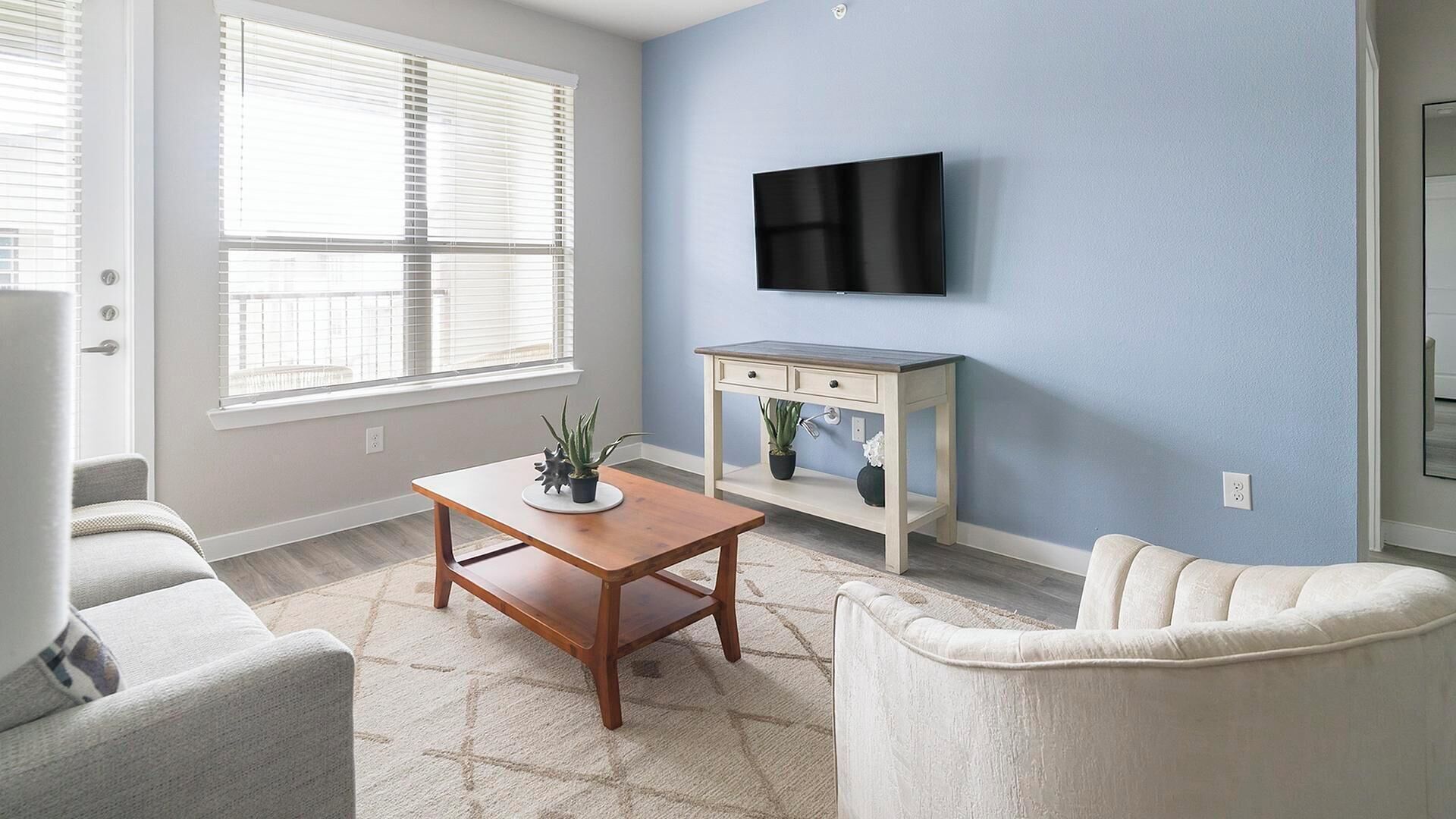 A living room with a wooden coffee table, two armchairs, a TV mounted on the wall, and a small white cabinet with plants.