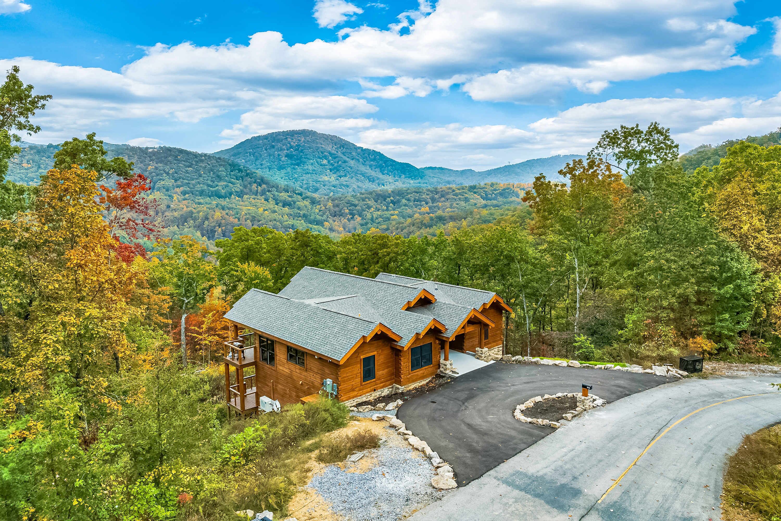 A wooden cabin with a curved driveway, surrounded by autumn foliage and mountains in the background.