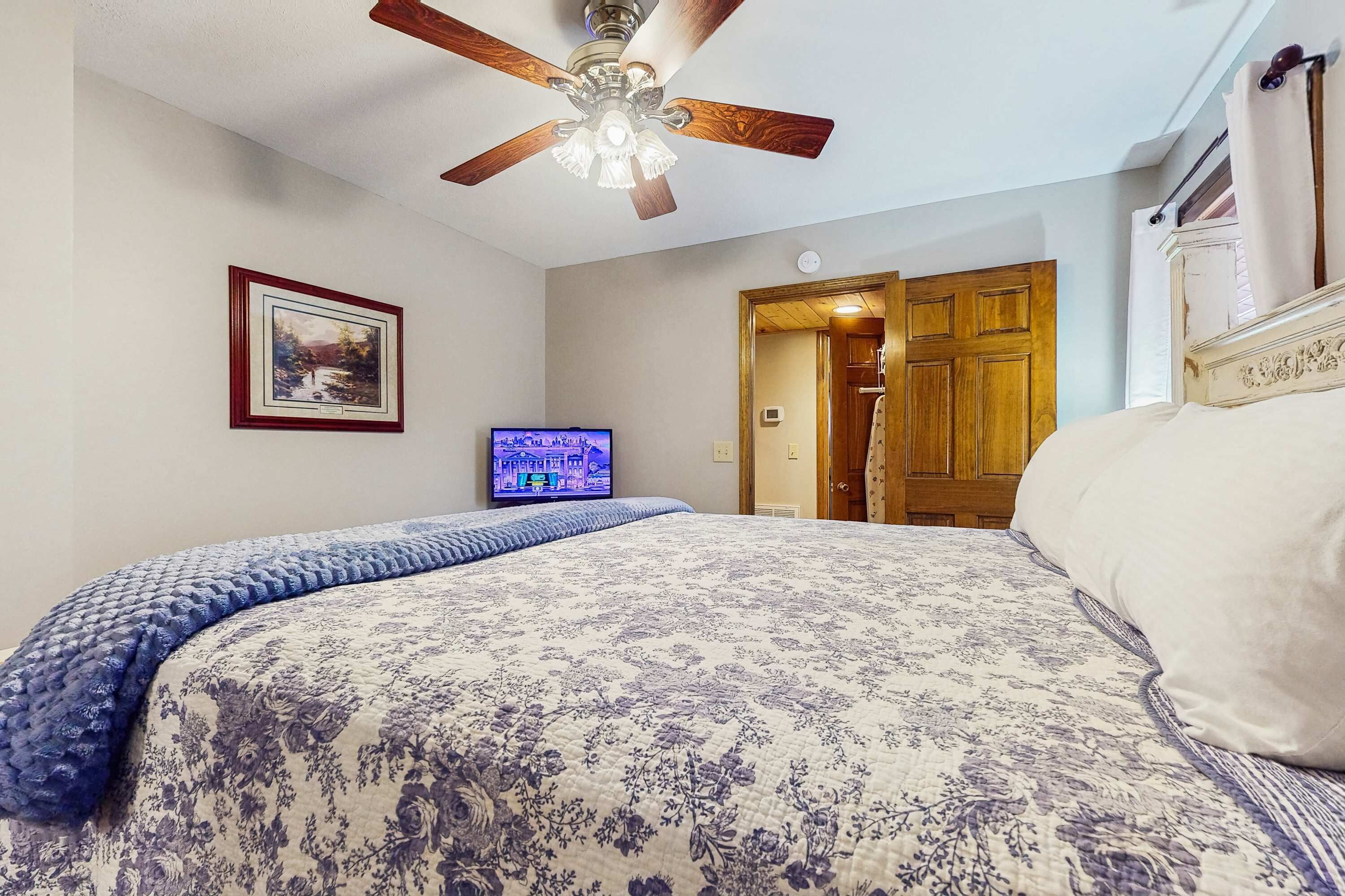 A bedroom with a bed, a ceiling fan, a television, and a door leading to another room.