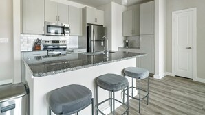 A modern kitchen with white cabinets, stainless steel appliances, a granite countertop, and bar stools. - Landing | Amazing 2BD, Gym, Pool (Katy)
