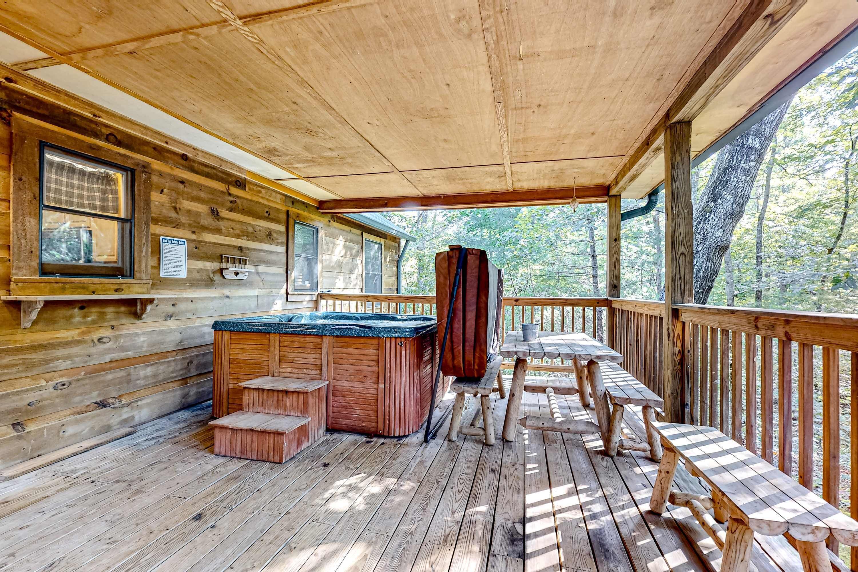 Una terraza de madera con jacuzzi, mesas de picnic y un área cubierta con ventanal a un bosque.