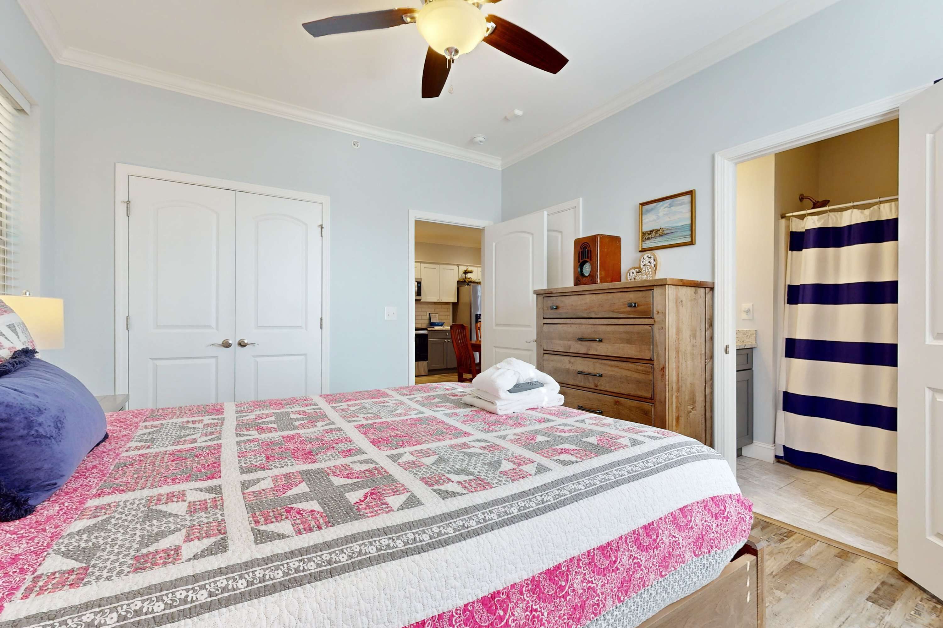 Une chambre à coucher comprenant un lit, un ventilateur de plafond, une commode et une salle de bain avec un rideau de douche à rayures.