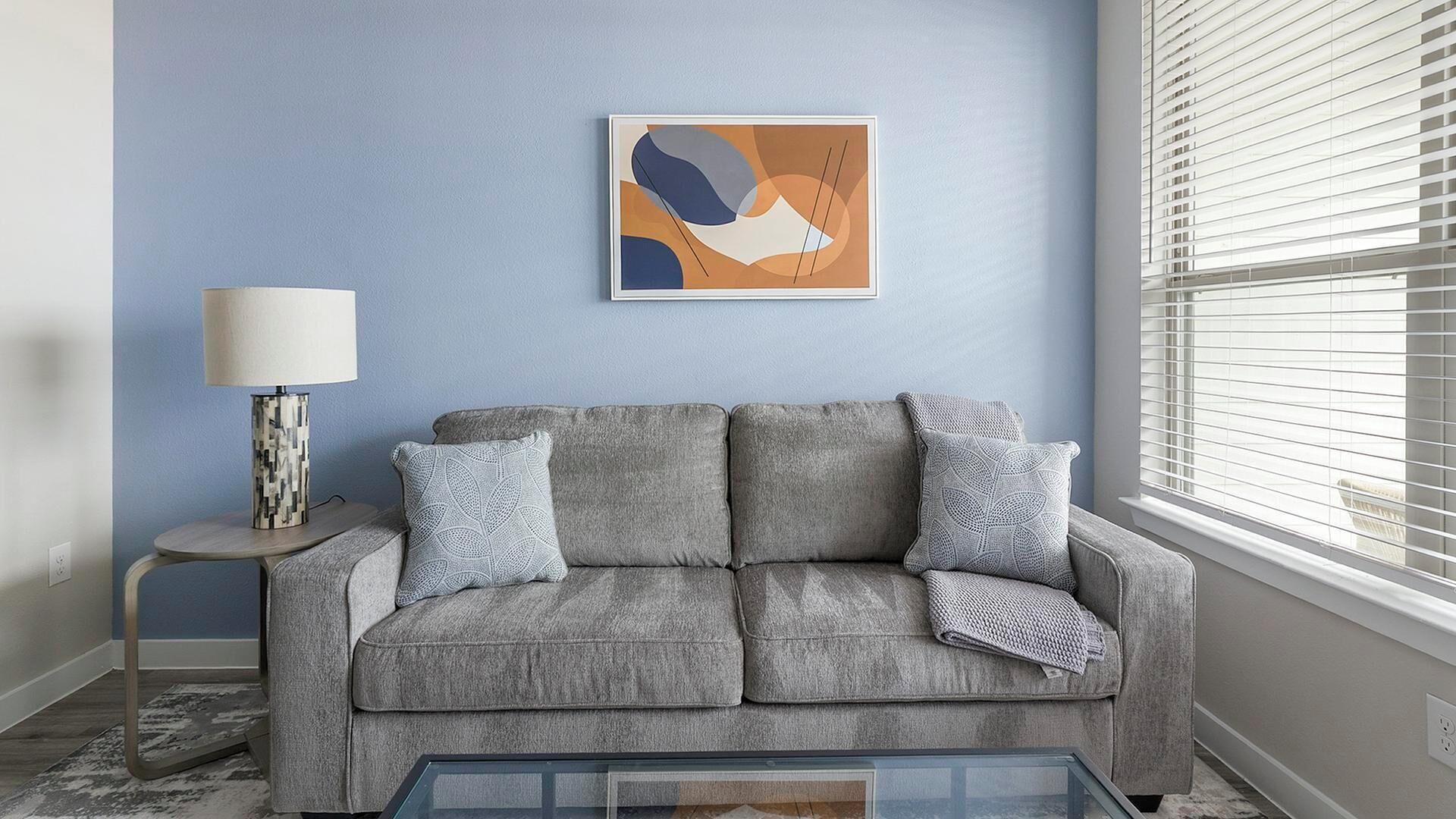 A grey sofa with patterned cushions, a side table with a lamp, and a framed abstract artwork on the wall.