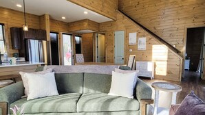 Interior acolhedor de uma cabana com um sofá verde, paredes de madeira, uma área de cozinha e uma escada.