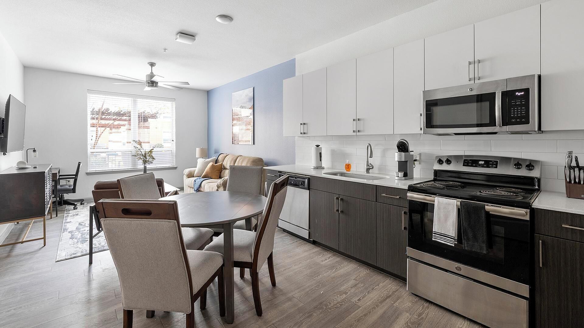A modern kitchen with a dining area, featuring stainless steel appliances, a dining table with chairs, and a view of the city through the window.