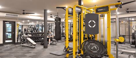 Une salle de sport équipée de divers appareils d’exercice, notamment une machine jaune pour l’entraînement musculaire, un banc et un sac de frappe.