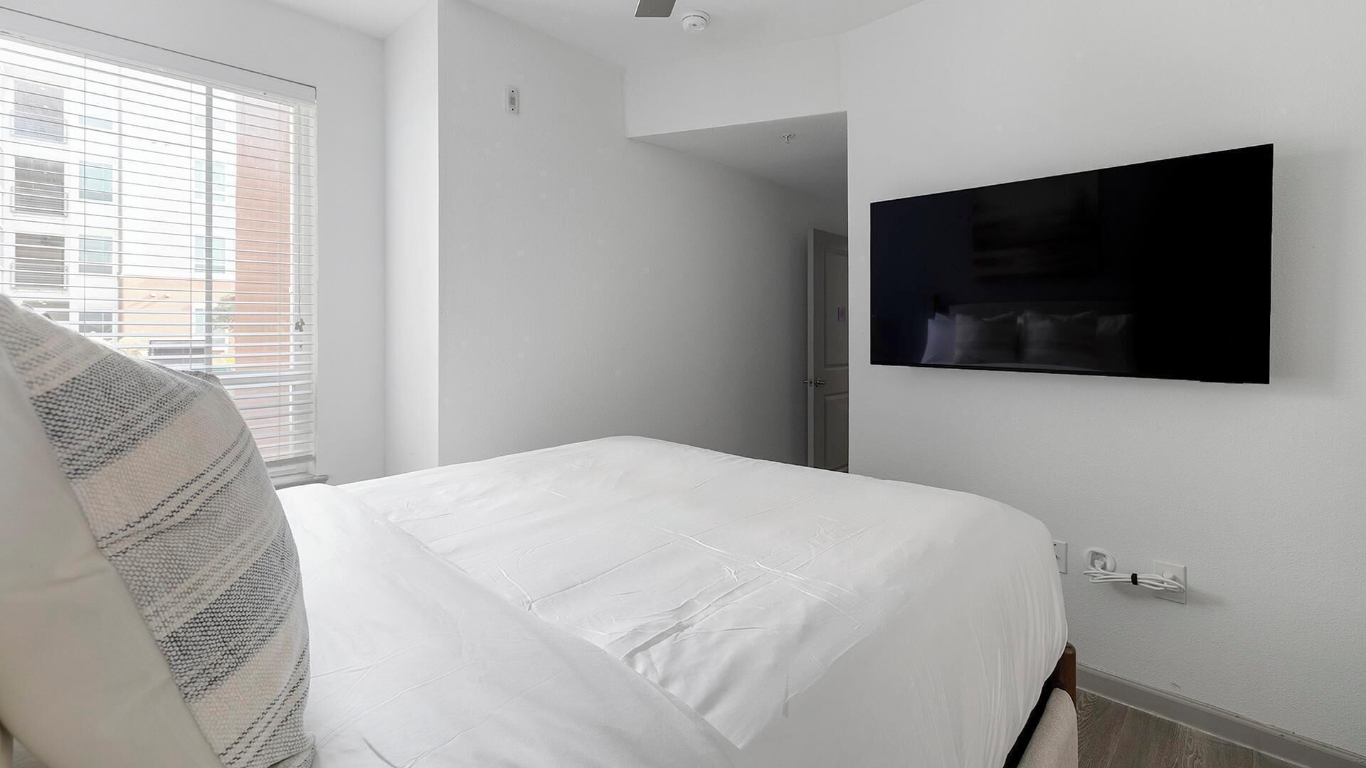 Een hotelkamer met een bed, een aan de muur bevestigde televisie en een raam met luxaflex.