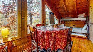 Una cabaña acogedora con interior de madera, una mesa redonda con mantel rojo, una cama litera y un ventanal con vistas a árboles otoñales.