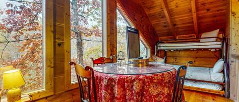 A cozy cabin with a wooden interior, a round table with a red tablecloth, a bunk bed, and a large window offering a view of autumn trees.
