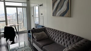 A modern living room with a grey tufted sofa, a glass coffee table, and a view of the city through large windows. - Iceboat Elegance Suites (Toronto)