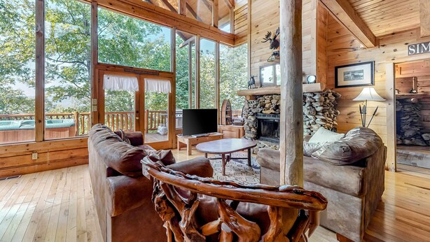 A wooden cabin interior with large windows, a stone fireplace, and a cozy seating area. - Charming Dog Friendly Cabin・Hot Tub・Scenic View (Sevierville)