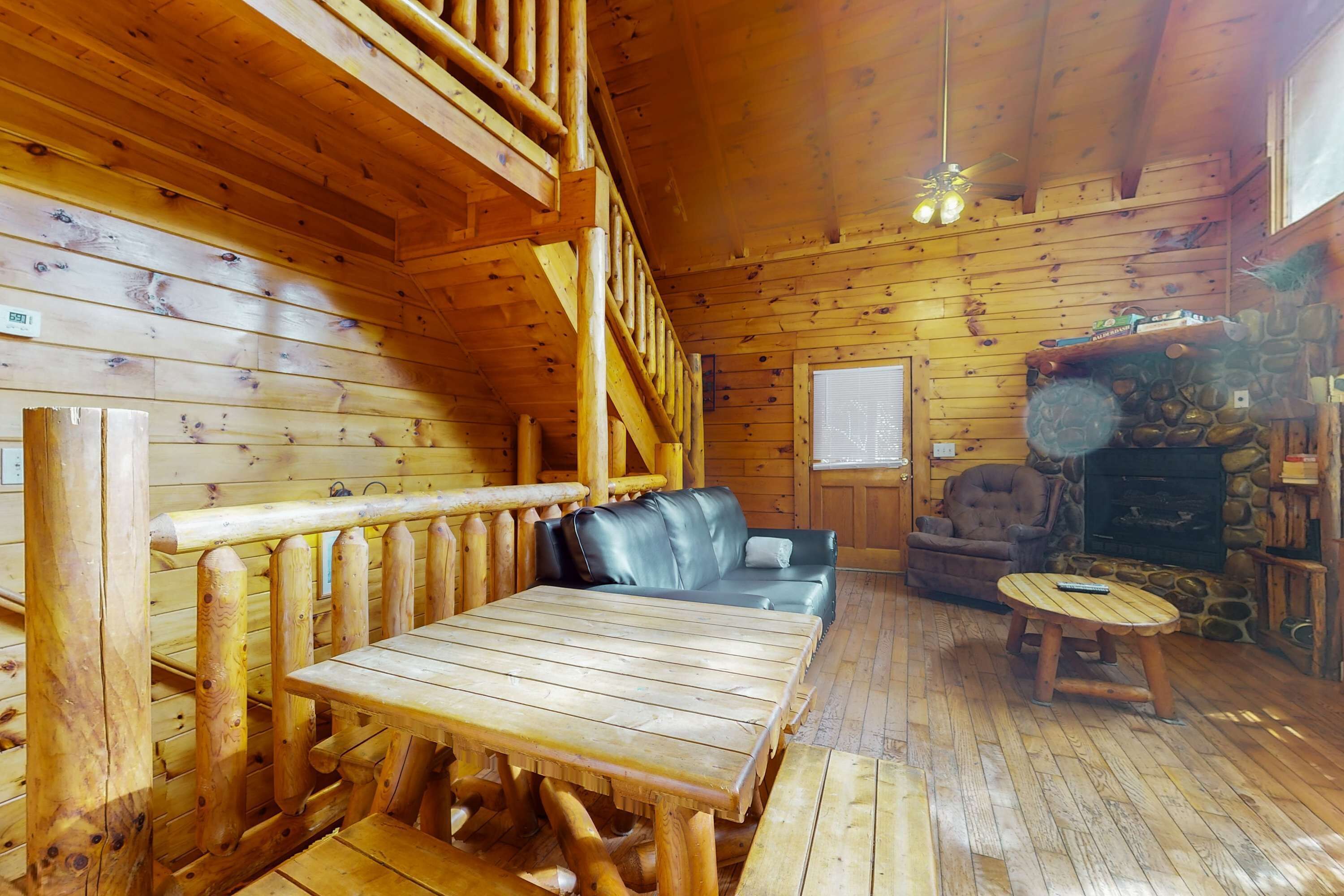 A log cabin interior with a wooden table, a leather sofa, a stone fireplace, and a wooden railing.