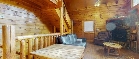 A log cabin interior with a wooden table, a leather sofa, a stone fireplace, and a wooden railing.