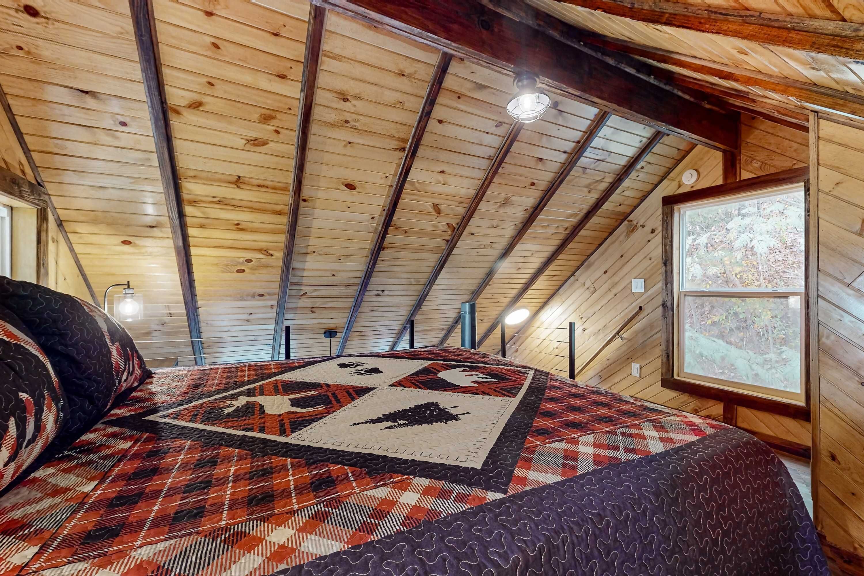 A cozy attic bedroom with a wooden ceiling, a quilted bedspread, and a window with a view of trees.