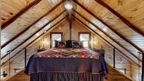 Une chambre avec un plafond en pente en bois, un lit recouvert d’une couvre-lit à motifs, deux fenêtres et une rambarde en bois le long du plafond.