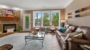 A living room with a stone fireplace, a glass coffee table, a leather sofa, and a view of trees through large windows.