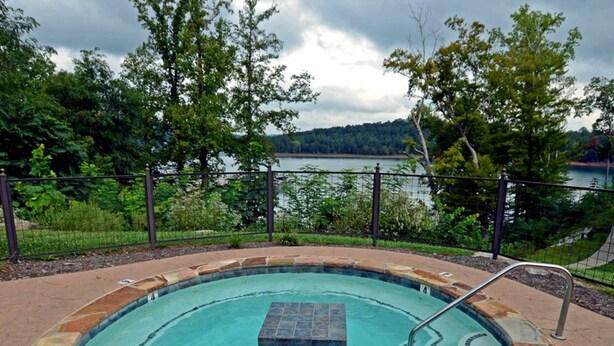 A swimming pool with a stone border and a metal ladder, surrounded by a fence and trees, with a lake and hills in the background.