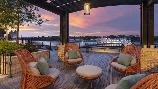A covered outdoor seating area with wicker chairs and a wooden deck, overlooking a river with a boat and cityscape in the background. - Landing | Chic 1BD, Clubhouse, Gym (Savannah)