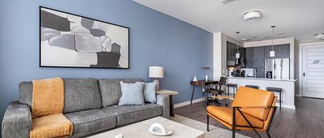 A modern living room with a grey sofa, orange armchair, and a coffee table. A kitchen is visible in the background with a white door.