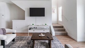 Un salon moderne avec un téléviseur à écran plat fixé au mur, un meuble de rangement blanc, une table basse en bois et un escalier menant à un autre niveau.