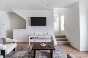 A modern living room with a flat-screen TV mounted on the wall, a white entertainment unit, a wooden coffee table, and a staircase leading to another level. - Landing | Spectacular 2BD, Clubhouse, Gym (Billings)