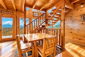 A wooden cabin interior with a dining table and chairs, a staircase, and a view of the mountains. - Spacious Family Friendly Getaway・Mtn View・Games (Pigeon Forge)