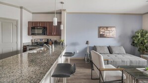 Una cocina moderna con encimeras de granito, microondas integrado y electrodomésticos de acero inoxidable. Cuenta con una zona de comedor con mesa y sillas de vidrio, y una sala de estar con sofá y una planta grande.