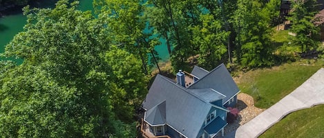 An aerial view of a house with a large deck, surrounded by lush green trees and a clear blue lake.