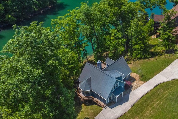 An aerial view of a house with a large deck, surrounded by lush green trees and a clear blue lake. - Waterfront Retreat, Sleeps 14, Theater, Games, Grill (Maynardville)